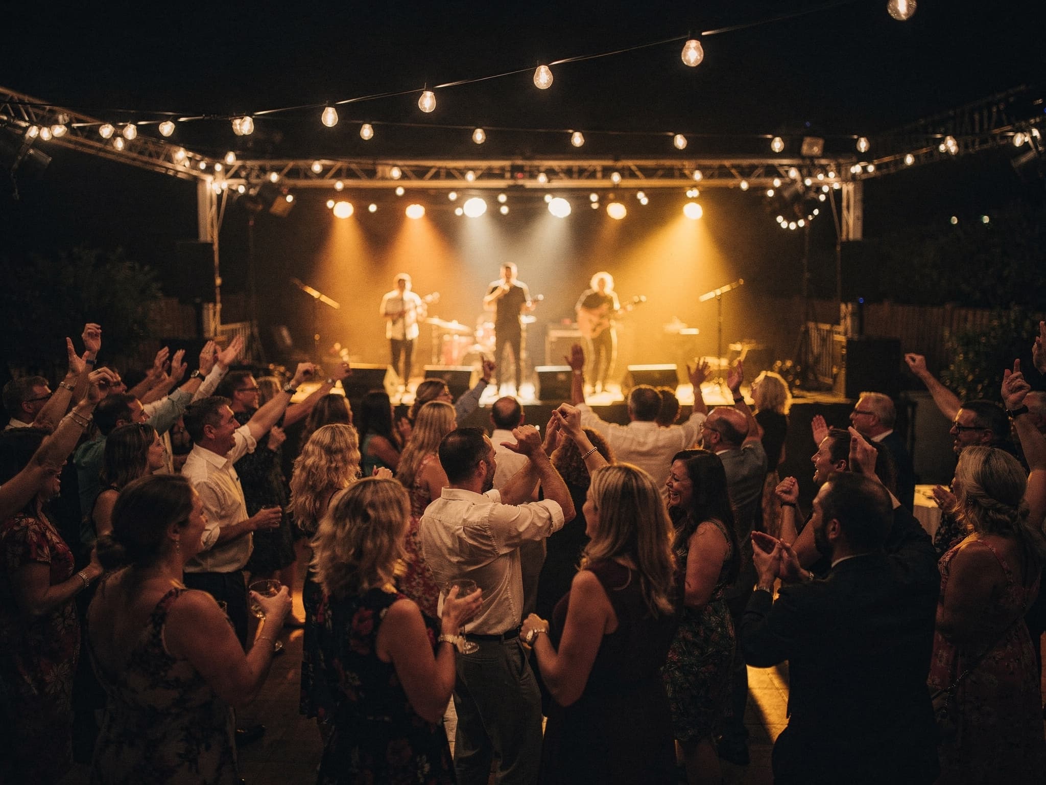 A packed wedding dance floor with the band playing on stage under string lights.